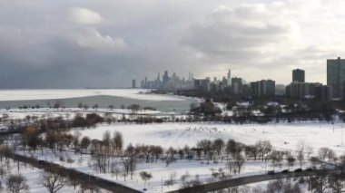 Panning out aerial drone clip of people sledding on Cricket hill surrounded by bare trees with a view of the downtown Chicago skyline and ice and snow on Lake Michigan.