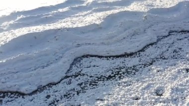 Aerial drone footage flying over a snow covered lake and beach with drifts and layers of ice and shadows covered in trails of human and animal footprints.