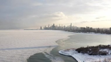 Panning out aerial of the Chicago skyline with cloudy sky above and snow and ice covering most of the water of Lake Michigan at sunset.