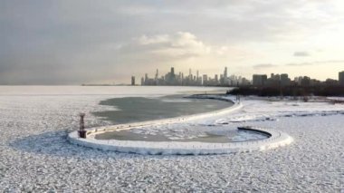 Panning in winter aerial cityscape scene flying towards a hooked shaped pier with snow encrusted ice chunks floating on Lake Michigan below and Chicago skyline in the distance at sunset. 