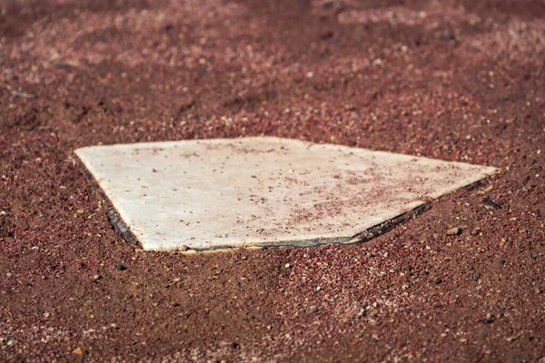 Close up of a white baseball home plate base in a field of wet brown or red colored dirt and mud on an empty baseball field on a sunny day.