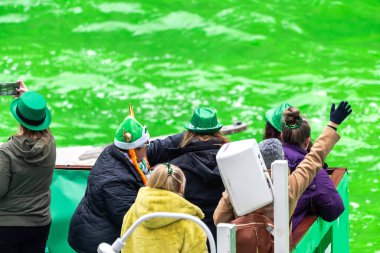 Chicago, IL - 11 Mart 2023: Aziz Patrick günü kostümleri ve yeşil şapkalarla bir tur teknesinde bekleyenler ve yıllık etkinlik sırasında nehri boyayan teknelerin fotoğraflarını çekenler.
