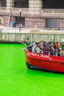 Chicago, IL - 11 Mart 2023: Parlak kırmızı Fred A. Otobüs teknesi, geleneksel St. Patrick nehri boyama etkinliğinin parlak yeşil suyuyla tezat oluşturan müşterileriyle nehir boyunca ilerliyor..