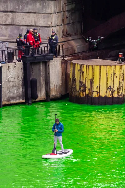 シカゴ 2023年3月11日 毎年恒例の聖パトリックの日イベント中に 新しく染められた緑の川の上のスタンドアップパドルボードに電話とパドルを持っている男の上に無人機が浮かんでいます — ストック写真