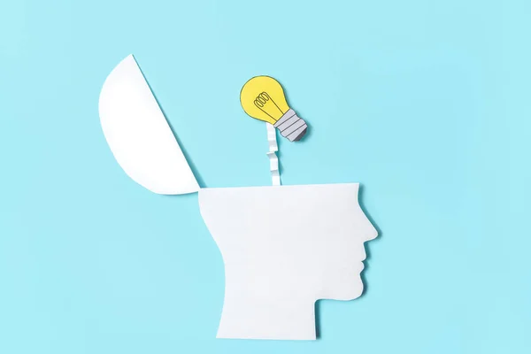 A close up of a cut out of a paper profile of a human head opening up with a light bulb popping out on a light blue background as a concept image for thinking of a creative idea.