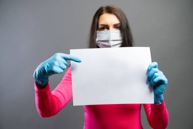 Tıbbi maskeli ve eldivenli genç bir kadın ellerinde boş kağıt olduğunu gösteriyor. Bir kağıt parçasına odaklan. Korona COVID-19 ve SARS cov 2 enfeksiyonlarını önler, her bir metin için boşluğu kopyala