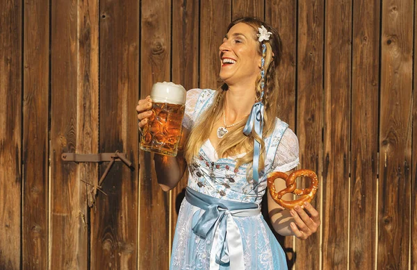 stock image Happy woman in traditional Bavarian Tracht holding a huge mug of beer, holding pretzel in other hand in Bavarian beer garden or oktoberfest