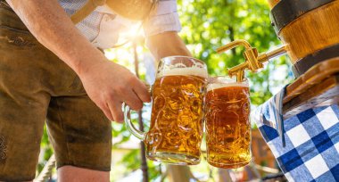 Deri pantolonlu Bavyeralı adam bira bahçesindeki ahşap bira fıçısından bira fıçısına bira dolduruyor. Oktoberfest, halk ve bira festivali için arka plan (Almanca: Ozapft)!) 