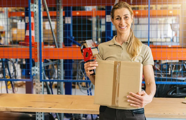 Happy female worker holding packing case after sealing it with adhesive tape at warehouse of freight forwarding company