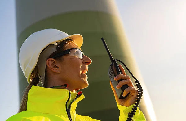 female engineer with safety jacket using walkie talkie to checking wind turbine system at windfarm