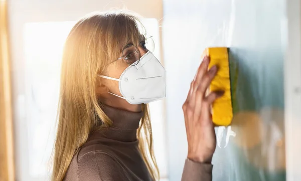 stock image teacher at schoool cleaning blackboard or chalkboard with sponge at class room and wearing protective ffp2 KN95 face mask due to coronavirus pandemic. 