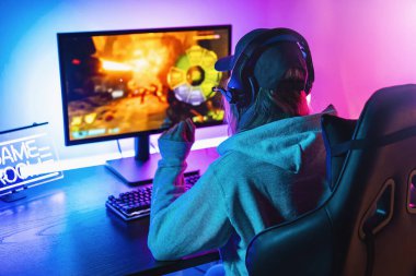 Kadın Oyuncu, Güçlü Kişisel Bilgisayarında Birinci Şahıs Shooter Online Video Oyunu oynuyor. Oda ve bilgisayar Renkli Neon Led Lights 'a sahip. Genç kadın evde şapka takıyor..