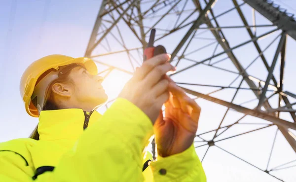 Electrical female engineer watching up and uses a walkie talkie to control a high voltage electricity pylon. Electrical power lines and towers with bright sun