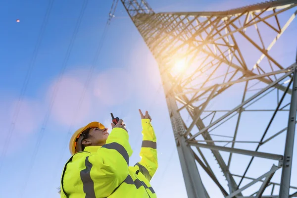 Electrical female engineer talking in a walkie talkie and pointing to control a high voltage electricity pylon. Close-up Electrical power lines and towers with bright sun