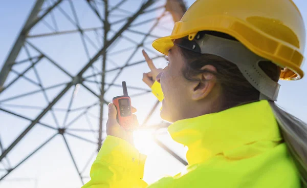 Electrical female engineer using walkie talkie to control a high voltage electricity pylon. Electrical power lines and towers with bright sun