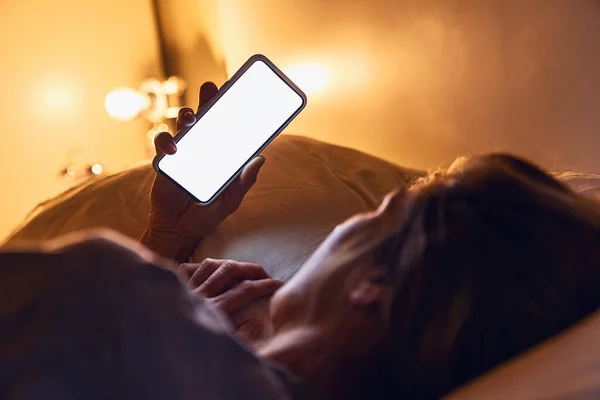 Woman holding smartphone late at night with white background for copyspace in bed. Person looking at blank phone app in dark home. Sexting or stalking concept image.