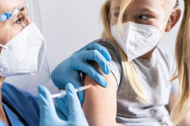 Doktorun ofisinde maske takan küçük bir kız aşı oluyor. Kovid 19 koronavirüs aşısı, grip, tehlikeli bulaşıcı hastalıklar için şırınga. İnsan ve çocuk için yapılan klinik denemelerden sonra iğne. 