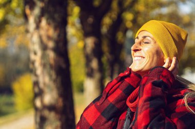 Kadın güneşli bir sonbahar gününün tadını çıkarıyor ve gülümsüyor. Renkli yapraklar ve ağaçlarla parkın ortasında duruyor ve dinleniyor. Temiz hava alıyor.