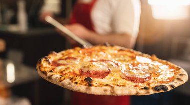 Restoran şefi fırından salamlı pizza alıyor. Geleneksel İtalyan pizzasını hazırlıyorum. Aşçı, odun ateşinde pişirilmiş İtalyan pizzasıyla kürek tutuyor..