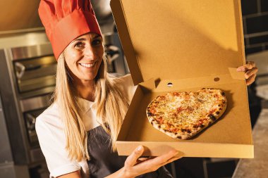 İtalyan pizzacıya teslim etmek için bir kutuda taş fırından kalp şeklinde taze peynirli pizza tutan kadın pizzacı.