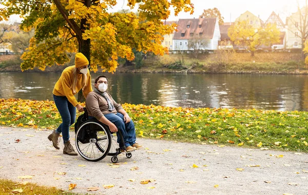 Woman and her friend with face mask in a wheelchair having stroll through the park at a river enjoying the autumn during covid-19 pandemic