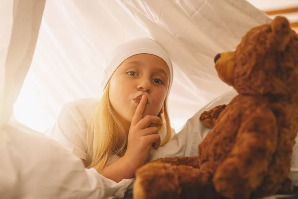 Sweet little girl with a teddy bear hold finger on lips showing silence to keep a secret while lies on her bed in a in tent with nightcap.