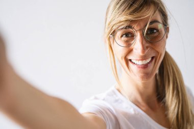 Sıradan giyinmiş çekici sarışın kadın ve gözlük geniş bir gülümseme. Güzel bir kadın beyaz arka planda poz verirken neşeli bir ifade takınıyor. Selfie için kollarını kameraya uzatıyor.
