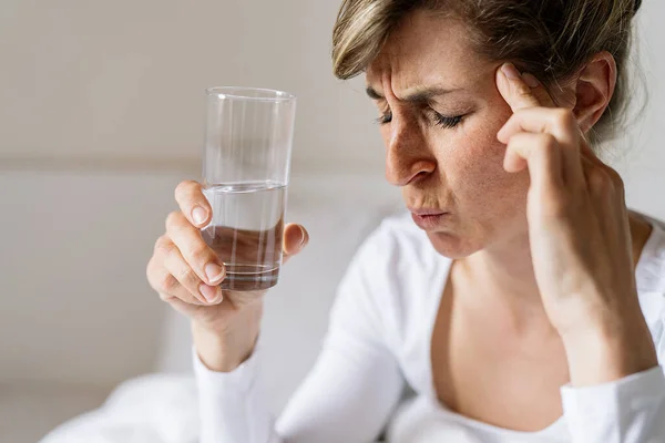 tired woman touching forehead and holding glass with water having headache migraine or depression, feel stressed grabs head with hand with pain sit on bed in the morning, Flu or overload Concept