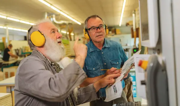Two men with ear protection discussing work on a cutting machine in a busy carpentry workshop