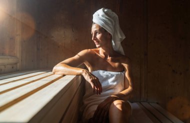 Finniş saunasında oturan bir kadın, bir koluna yaslanmış, kafasına bir havlu sarılmış, sağlık spa otelinde güneş ışığı yayılıyor.