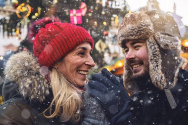 Çiftler el ele tutuşurken sıcak bir anı paylaşıyorlar. Arka planda neşeli bir Noel pazarı var.