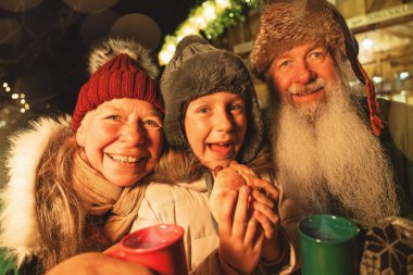 Torunu olan mutlu büyükanne ve büyükbaba sosisli sandviç yiyor ve Noel pazarında sıcak şarap bardakları tutuyor.