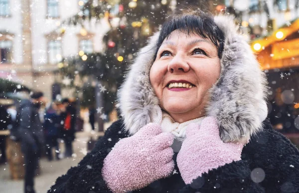 Elderly woman smiling at a winter Christmas market, with snow falling, in festive attire