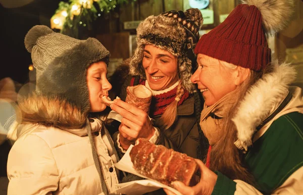 Happy Grandma and mother feed their daughter with a Baumstriezel or trdelnik in front of a Christmas market in Germany
