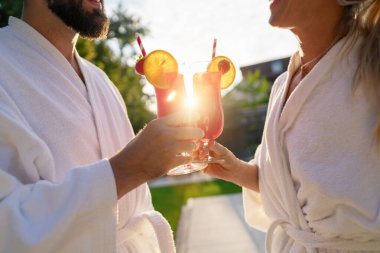 Couple in white bathrobes clinking red cocktails with a sunset behind at spa hotel clipart