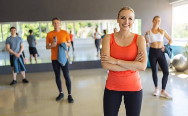 Spor salonunda kollarını kavuşturmuş turuncu kollu tişörtlü gülümseyen bir kadın. Arka planda insanlar var. Takım çalışması Görüntü kavramı