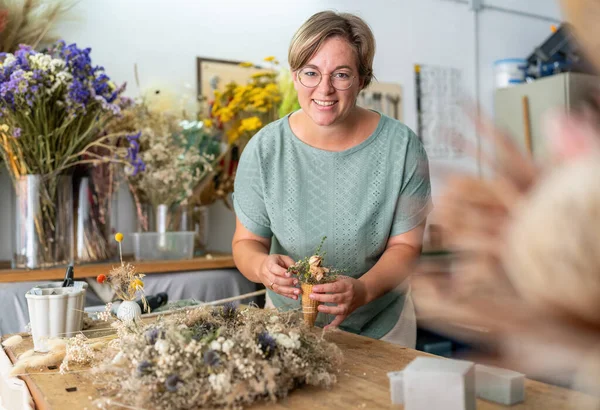 cheerful woman in a mint-green blouse delicately arranges dried flowers inside a cone, surrounded by a vibrant studio setting filled with diverse flora