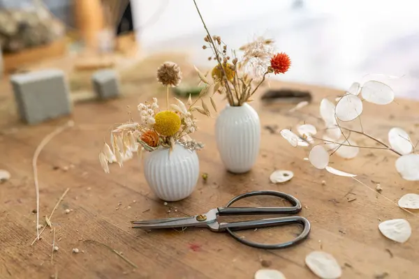 Two white ribbed vases with dried flowers on a wooden table with scattered petals and scissors