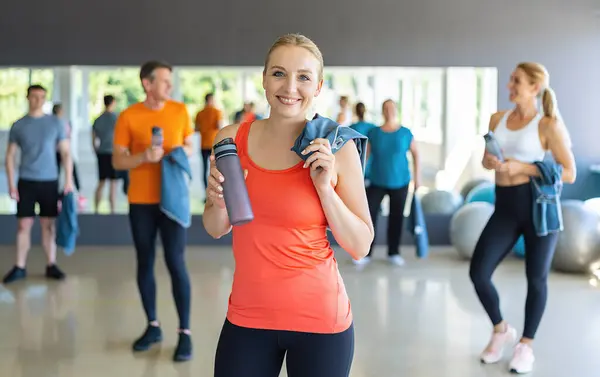 Woman in orange top holding a water bottle in a gym with other people in the background. Teamwork Concept image