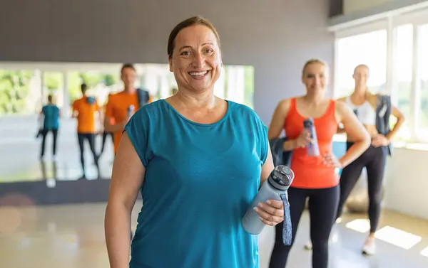 Smiling older woman holding a water bottle in a gym with other participants in the background. Teamwork Concept image