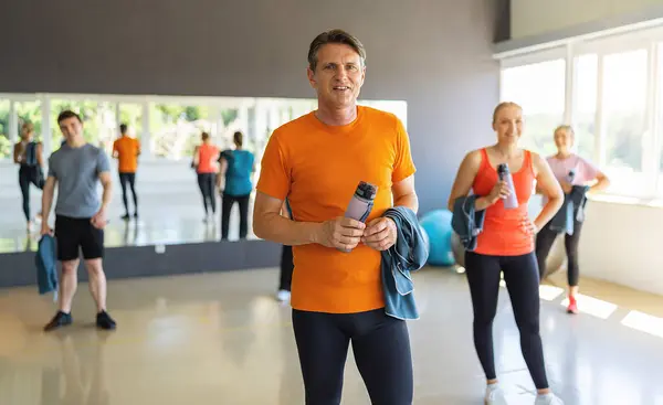 Man in orange shirt holding a water bottle in gym with other people and exercise balls. Teamwork Concept image