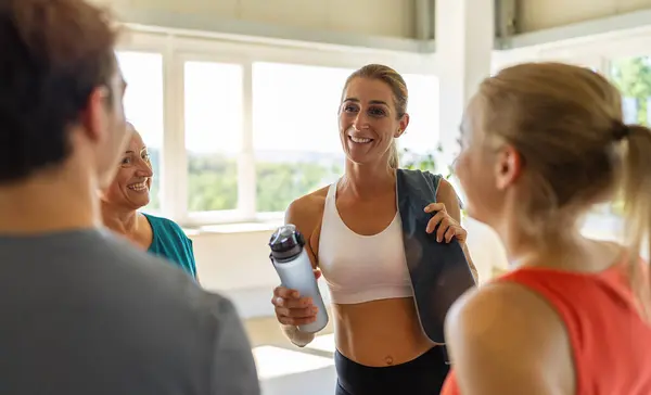 Smiling people chatting in a gym with a woman holding a water bottle in the foreground. Teamwork Concept image