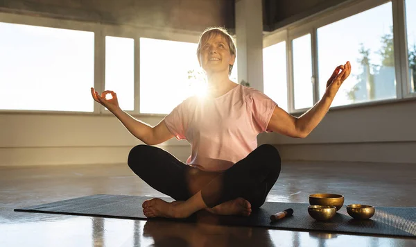 Woman meditating in gym with singing bowls, sunlight, peaceful yoga practice