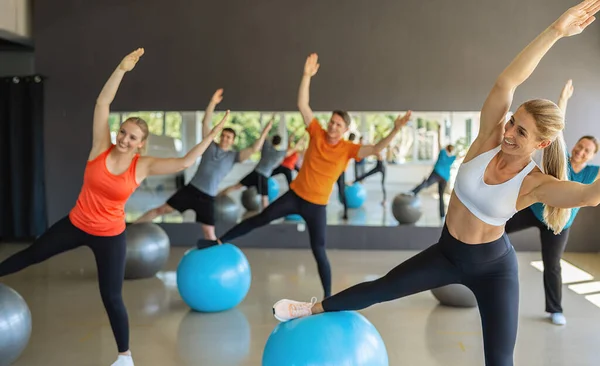 Group of happy people in a receding line doing pilates in a gym balancing on a gym balls with hands up toning their muscles. Healthy sports lifestyle, Fitness, Healthy concept.