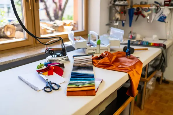 sewing or tailoring workshop showcases a table filled with sewing essentials: fabric swatches, scissors, iron, color palette, silk fabrics and tools, with a window displaying stacked firewood outside