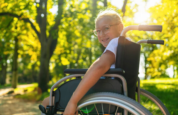 Smiling young handicapped blond girl sit in wheelchair during walk in park, teenager disability concept image