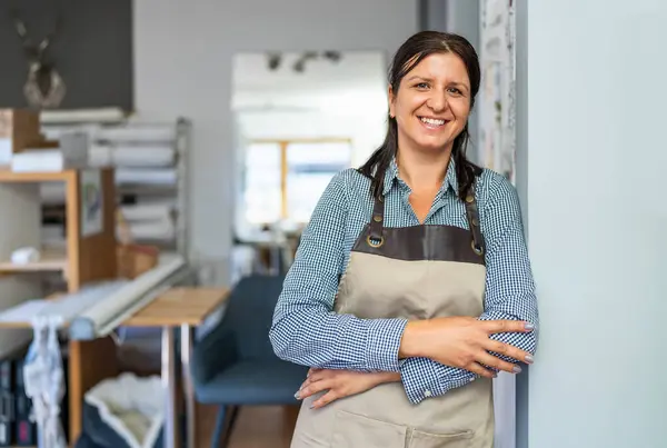 confident woman wearing a checkered shirt and beige apron stands in a workshop with folded arms, smiling towards the camera.