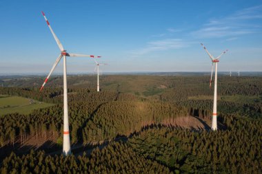Rüzgâr türbinleri, akşam güneşinin yansıttığı gölgelerle dolu ormanlık bir arazide yükseliyor. Yenilenebilir enerji konsepti