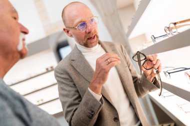 Optimetrist optik dükkanındaki müşteriye gözlük sunuyor. Gözlük rafının önünde gözlük seçimi sunuyor.