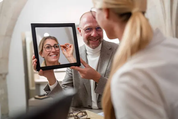 Optician holding a mirror to smiling woman trying on new glasses in a optical store.